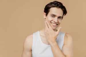 Attractive smiling young man in his 20s with perfect skin, touching his face after shaving, representing skincare, facial rejuvenation, and bio-care concepts
