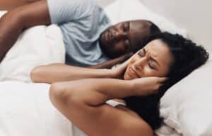 Frustrated woman covering her ears in bed while her partner snores, highlighting the benefits of NightLase® treatment for sleep apnea relief